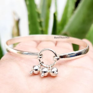 May include: A silver bangle bracelet featuring three small, dangling bell charms. The bracelet has a simple, polished design with a circular charm holder. The text "BaniThani925" is visible on the bracelet.