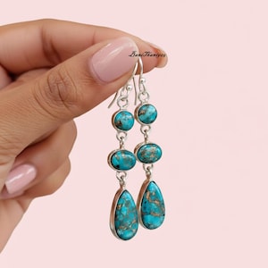 Puede incluir: Pendientes colgantes de plata con piedras turquesas. Cada pendiente tiene tres piedras turquesas en forma redonda, ovalada y de lágrima. Las piedras turquesas son de color azul verdoso con vetas marrones. Los pendientes son sostenidos por una mano.