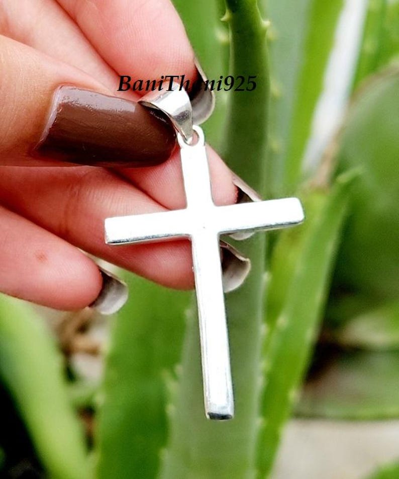 Pode incluir: Um pingente de cruz de prata com um design simples e cl&aacute;ssico. A cruz &eacute; segurada contra um fundo verde desfocado. O texto "BaniThani925" &eacute; vis&iacute;vel no canto superior esquerdo.