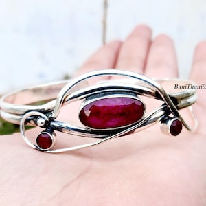 Peut inclure: Un bracelet en argent orné d'un grand rubis ovale au centre et de deux petits rubis. Le bracelet présente un design fluide et unique. Le texte "BaniThani925" est visible.
