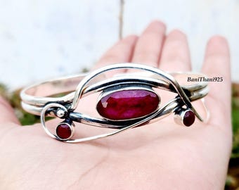 May include: A silver bracelet featuring a large oval ruby gemstone and two smaller ruby accents. The bracelet has a distinctive, flowing design. The text "BaniThani925" is visible on the bracelet.