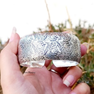 May include: A silver cuff bracelet with an intricate paisley pattern. The bracelet is wide and has a rounded shape.