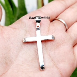 Pode incluir: Um pingente de cruz de prata polida. A cruz &eacute; segurada em uma m&atilde;o, com o texto "BaniThani925" vis&iacute;vel. O pingente &eacute; um s&iacute;mbolo religioso.