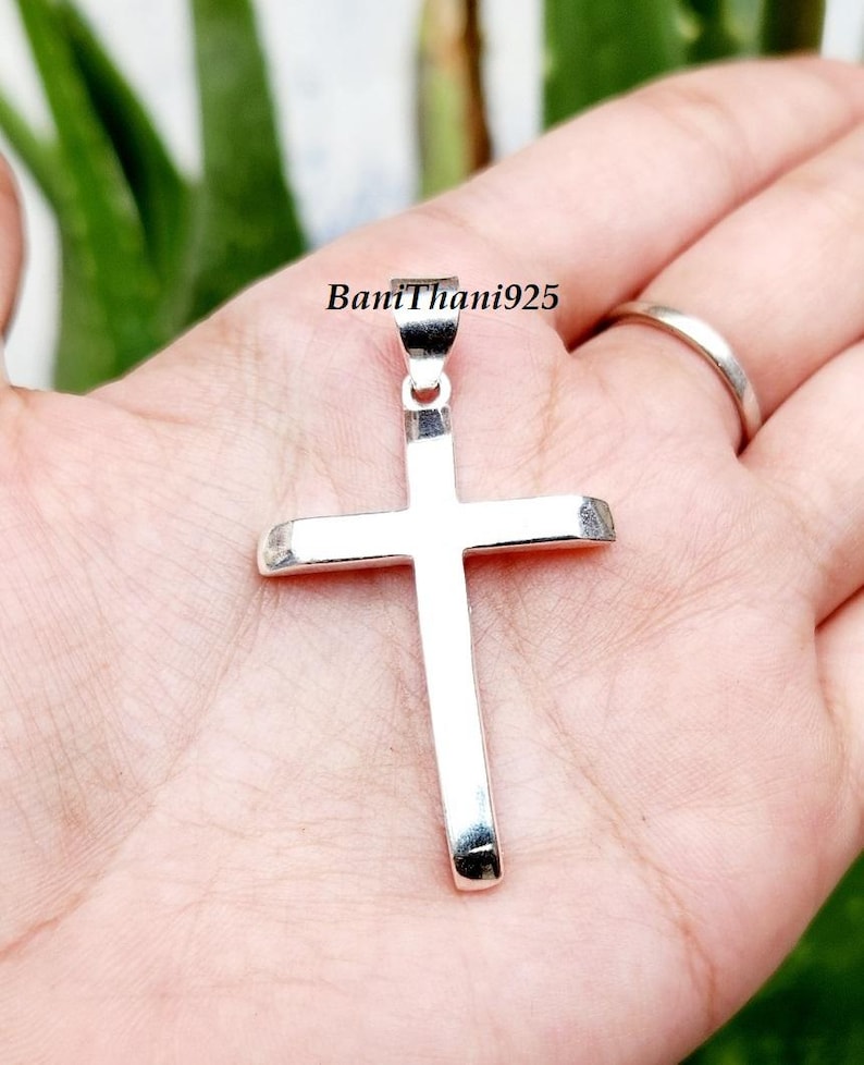 Pode incluir: Um pingente de cruz de prata polida. A cruz &eacute; segurada em uma m&atilde;o, com o texto "BaniThani925" vis&iacute;vel. O pingente &eacute; um s&iacute;mbolo religioso.