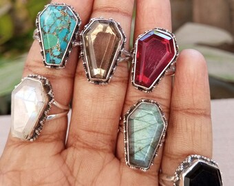 Puede incluir: Un conjunto de seis anillos de plata con piedras preciosas de diferentes colores. Cada anillo presenta un engaste en forma de ataúd. Las piedras preciosas incluyen turquesa, marrón, rojo, blanco, verde y negro.
