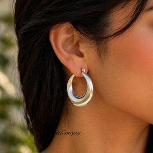 May include: A close-up of a silver hoop earring. The earring is a thick, polished, crescent shape. The earring is attached to the earlobe. The background is blurred.