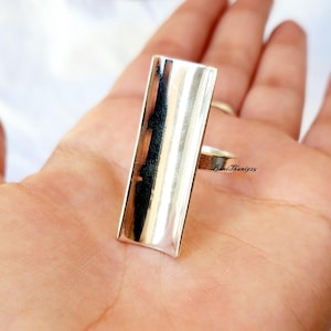 May include: A silver ring with a rectangular face. The ring is highly polished and reflects light. The band is thin and simple. The ring is shown on a hand.