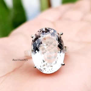 May include: A clear, oval-cut gemstone ring with a silver band. The large, faceted stone reflects light, showcasing its clarity. The ring is set on a light-coloured surface, with the text "BaniThani925" visible.