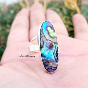 May include: A silver ring featuring an oval abalone shell inlay. The shell displays iridescent blue, green, and purple hues.