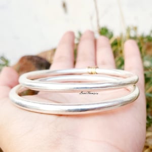 May include: Two silver bangles stacked on top of each other. The bangles are smooth and shiny, and the top bangle has a small gold accent.