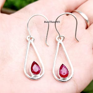 May include: A pair of silver drop earrings with a teardrop design. Each earring features a vibrant, red gemstone set within a silver frame. The earrings have a hook closure. The text "BaniThan925" is visible.