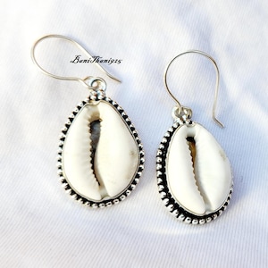 May include: A pair of silver earrings featuring teardrop-shaped white seashells. The shells are set in a beaded silver frame and hang from silver ear wires. The earrings are displayed on a white surface. The text "BaniThani925" is visible.