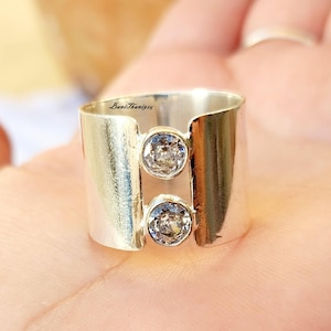 Puede incluir: Un anillo de plata con dos piedras transparentes engastadas en una banda ancha. El anillo está abierto por un lado y tiene una forma ligeramente curvada.