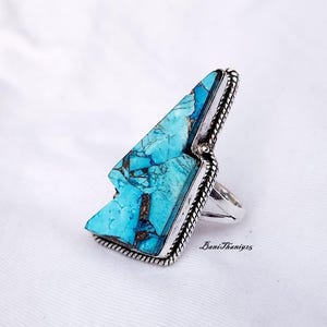 Pode incluir: Um anel de prata com uma grande pedra turquesa engastada em uma forma geométrica. A pedra tem uma cor azul e verde com veios marrons.