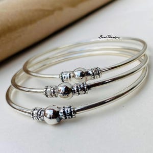 Op de afbeelding: Drie zilveren armbanden met een getextureerd ontwerp en een ronde kraal op elk. De armbanden zijn op elkaar gestapeld.