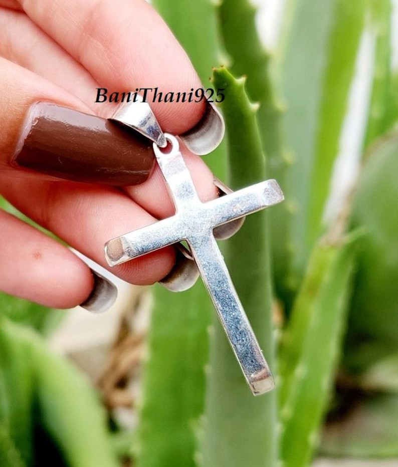 Pode incluir: Pingente de cruz prateada com um design simples e elegante. A cruz &eacute; segurada contra um fundo de plantas verdes. O texto "BaniThani925" &eacute; vis&iacute;vel na imagem.
