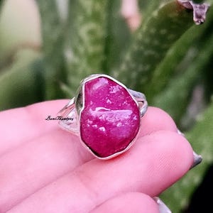 May include: A silver ring featuring a large, irregular-shaped, vibrant pink gemstone. The ring's band is a simple, polished silver, and the gemstone is the focal point. The ring is shown against a blurred green background.