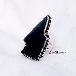 Pode incluir: Um anel de prata com uma pedra preciosa preta em forma de triângulo. O anel tem um design trançado ao redor da borda.