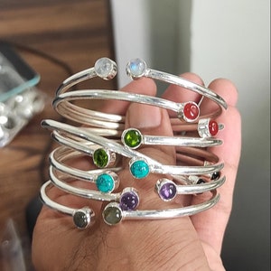 May include: A set of silver bangles with colorful gemstones. The bangles are stacked on top of each other and feature a variety of stones, including turquoise, amethyst, and green.