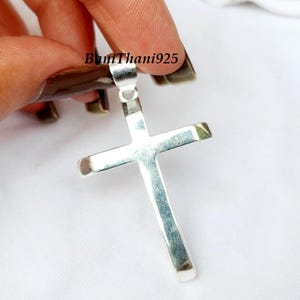 Pode incluir: Um pingente de prata em forma de cruz, segurado por uma m&atilde;o contra um fundo branco. O texto "BumThani925" &eacute; vis&iacute;vel na imagem.