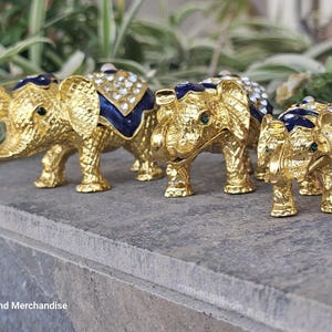 May include: A collection of gold-colored elephant figurines. Each elephant has a textured surface, adorned with blue enamel accents and sparkling rhinestones. The elephants are arranged on a stone surface, with greenery in the background.
