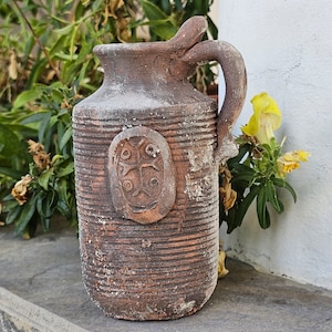 May include: A brown clay pitcher with a textured surface and a decorative design. The pitcher has a wide mouth and a handle.