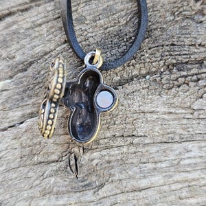 May include: A detailed close-up of a pendant necklace with a cross-shaped locket. The locket is open, revealing a small interior space. The pendant has a bronze finish with intricate detailing and hangs from a black leather cord.