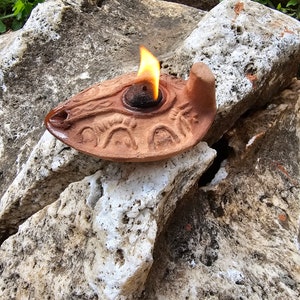 Può includere: Una lampada ad olio in terracotta con una fiamma che brucia all'interno. La lampada è decorata con un motivo di cerchi e linee. È appoggiata su una roccia.