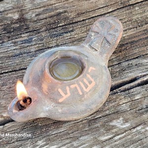 Puede incluir: Una lámpara de aceite de arcilla color canela con una mecha encendida, que presenta un depósito circular central, un asa con una cruz en relieve y una inscripción en hebreo. La lámpara tiene una textura terrosa y una pequeña llama cálida.