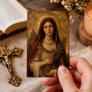 Op de afbeelding: Een rechthoekige gebedskaart met een klassieke illustratie van een figuur met een gouden aureool, gekleed in een donkerrode hoofddoek en een crèmekleurig gewaad, die een kleine harp en een groene palmtak vasthoudt tegen een textuur.