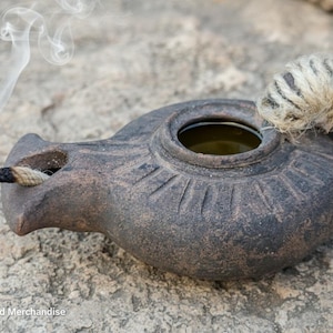 May include: A lit, antique-style oil lamp made of dark brown ceramic. The lamp has a wick, a flame, and a decorative rope handle. Smoke rises from the burning wick, and the lamp sits on a stone surface.