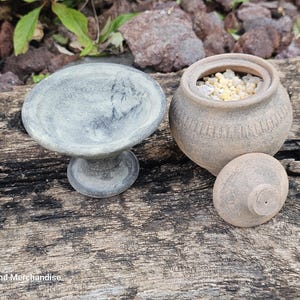 Pode incluir: Uma taça de pedra cinza com pedestal e um pequeno pote bege redondo com tampa. O pote contém pequenos grânulos de cor clara. Os itens estão dispostos em uma superfície de madeira envelhecida, sugerindo um tema natural ou rústico.