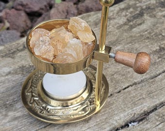 May include: A gold metal incense burner with a wooden handle. The burner has a round bowl filled with amber-colored resin. The burner is sitting on a wooden surface.