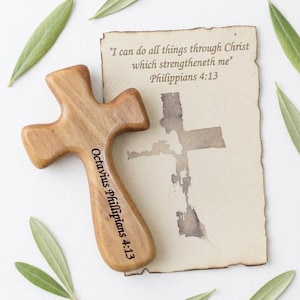 Könnte beinhalten: Ein Holzkreuz mit abgerundeten Kanten, graviert mit "Octavius Phillipians 4:13". Ein Pergamentstück zeigt den Text "Ich vermag alles durch den, der mich stark macht" Philipper 4:13. Grüne Blätter sind um das Kreuz und das Pergament verstreut.