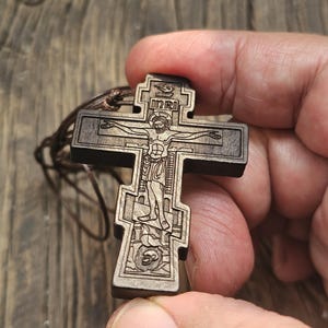 Puede incluir: Un colgante de cruz de madera marrón oscuro con intrincadas tallas de una figura religiosa y las letras INRI. La cruz está suspendida de un cordón marrón. Las palabras "Holy Land Merchandise" están en la parte inferior.