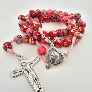 Puede incluir: Un rosario rojo y rosa con cuentas con estampado floral y un crucifijo de plata y un dije de corazón con una imagen de la Virgen María.