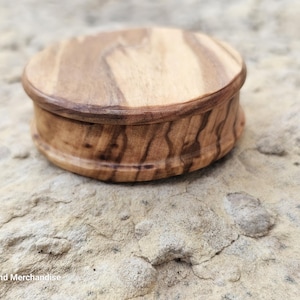 May include: A small, round wooden box with a lid. The box is made of light brown wood with darker brown grain patterns. The box is sitting on a rough, light-colored stone surface. The box appears to be handcrafted.