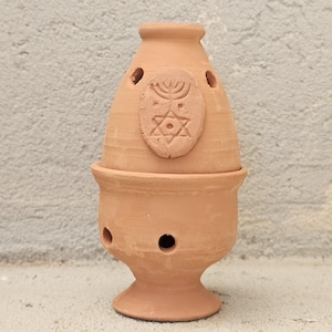 May include: A terracotta incense burner with a two-piece design, featuring a conical top and a pedestal base. The top section displays an embossed menorah and Star of David emblem. Both pieces have small circular ventilation holes throughout.