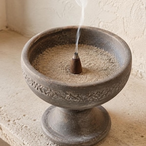 Pode incluir: Um queimador de incenso de cerâmica cinza-escuro em forma de pedestal com um topo largo em forma de taça com um padrão de faixa em relevo. A taça está cheia de cinzas cinza-claras e um cone de incenso castanho-escuro emite fumo no centro.
