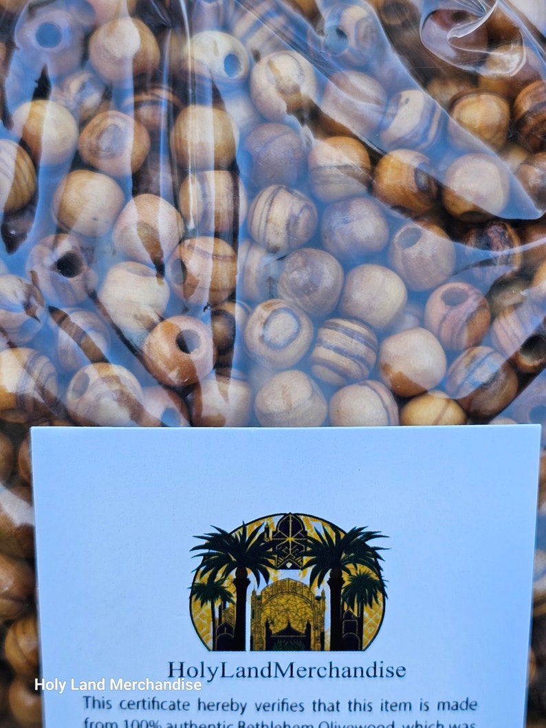 May include: A close-up of a bag of olive wood beads. The beads are a light brown color with dark brown stripes. The bag is clear plastic and has a certificate that says "HolyLandMerchandise" and "This certificate hereby verifies that this item is made from 100% authentic Bethlehem Olivewood which was..."