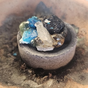 May include: A small, gray stone bowl filled with colorful, irregularly shaped pieces of blue, clear, and black materials. The bowl sits inside a larger, light brown, clay-like container. The text "Holy Land Merchandise" is visible at the bottom.