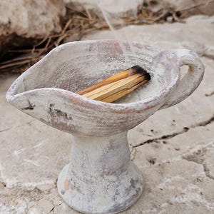 Pode incluir: Um queimador de incenso de argila moldado à mão com um acabamento rústico em tom esbranquiçado e uma alça lateral. O recipiente em forma de taça apresenta um bico largo e contém um pedaço de madeira de palo santo fumegante com fumaça.