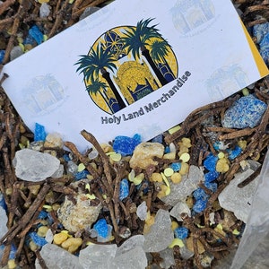 Könnte beinhalten: Nahaufnahme eines Beutels, gefüllt mit einer Mischung aus braunen Stöcken, klaren Glasstücken, blauen und gelben Perlen. Eine weiße Karte mit einem Logo und dem Text "Holy Land Merchandise" ist oben auf der Mischung sichtbar.