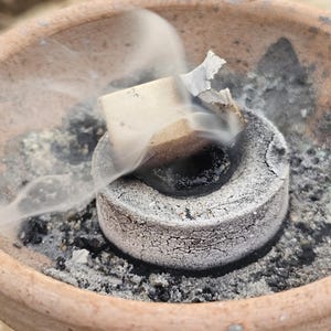 May include: A terracotta bowl containing a lit charcoal disc, with smoke rising. A small, square incense piece is burning on top of the charcoal. The image is labelled "Holy Land Merchandise".