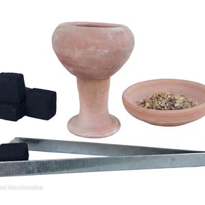 Puede incluir: Un quemador de incienso de terracota con base de pedestal, un pequeño cuenco de terracota con incienso en resina, tres cubos de carbón negro y unas pinzas de metal plateado que sostienen un cuarto cubo de carbón. El texto Holy Land Merchandise es visible.