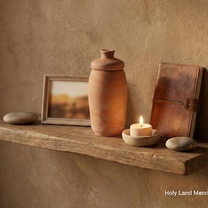 Puede incluir: Una estantería rústica de madera exhibe un jarrón de terracota con tapa, un marco con foto, un diario de cuero, una vela encendida y piedras lisas. La estantería está montada en una pared marrón texturizada, creando un ambiente cálido.
