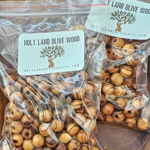 May include: Two bags of olive wood beads. The bags are clear plastic and have a label that says "Holy Land Olive Wood" and "HolyLandMerchandise.com". The beads are a light brown color with dark brown stripes.
