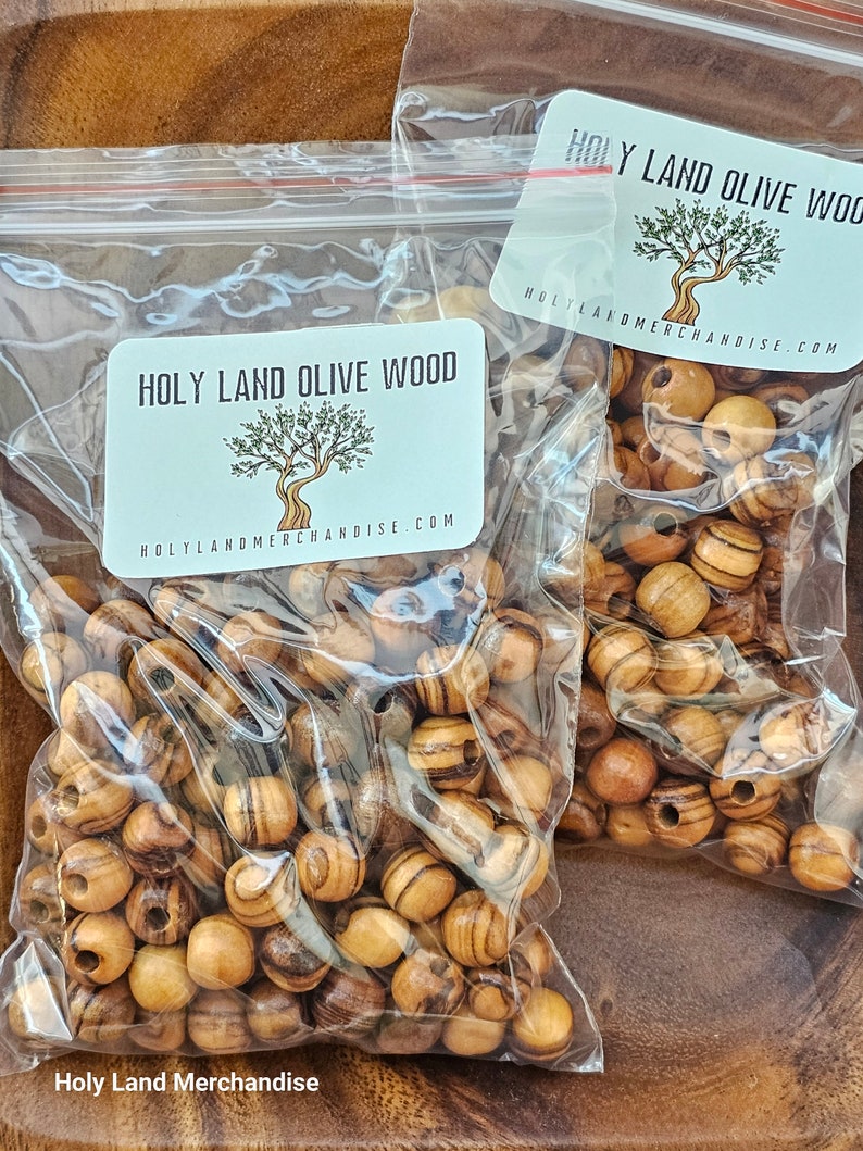 May include: Two bags of olive wood beads. The bags are clear plastic and have a label that says "Holy Land Olive Wood" and "HolyLandMerchandise.com". The beads are a light brown color with dark brown stripes.