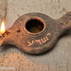 May include: A brown clay oil lamp with a lit wick and a central circular opening. The surface features textured, weathered details and Hebrew characters painted in a light orange hue. The lamp has a handle on one side and a spout for the flame.