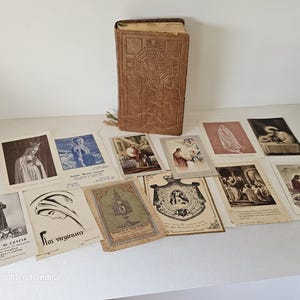 以下が含まれることがあります： 茶色の革張りの本と、エンボス加工された十字架のデザイン、ヴィンテージの祈りのカードなど、宗教的なアイテムのコレクション。カードには宗教的な人物やシーンが描かれており、テキストも表示されています。白い表面に配置されています。